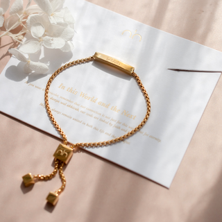 Gold bracelet with charms on a white card with text, placed on a light pink surface.